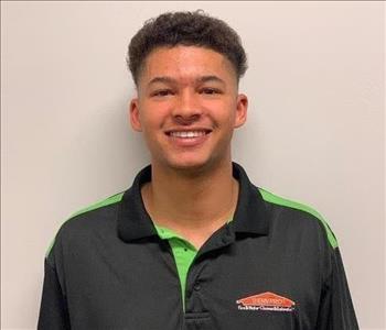 Male with black curly hair wearing a black shirt with SERVPRO on it, in front of a grn/gry wall.